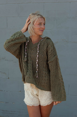A woman wearing an oversized chunky knit pullover sweater in olive green with braided drawstrings, paired with cream lace shorts, standing against a gray painted brick wall.