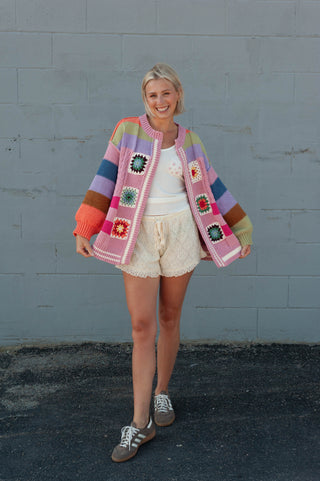 A woman modeling a colorful striped crochet cardigan with granny square patches, paired with cream shorts and sneakers, standing against a gray painted brick wall.
