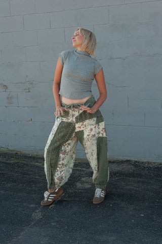 A woman wearing a gray short-sleeve crop top and olive green patchwork harem pants with a floral print, standing against a gray concrete block wall.