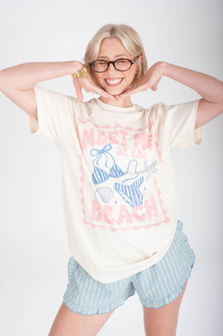 Person wearing a graphic t-shirt with 'Meet Me at the Beach' text and blue shorts on a white background