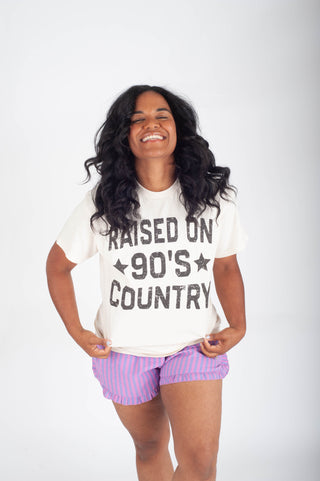 Woman wearing a 'Raised on 90's Country' t-shirt and purple shorts on a white background