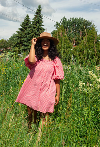 Woman in a pink dress and hat standing in a field of tall grass and wildflowers.