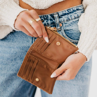 A close-up of a person holding an open brown leather wallet or card holder with gold-tone hardware, wearing blue jeans and a cream knit sweater.