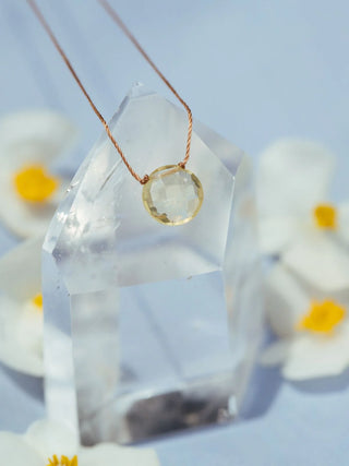 Necklace with a gold pendant on a clear crystal prism against a light blue background with flowers.