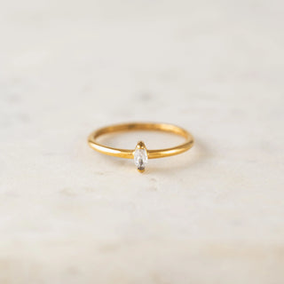 A delicate gold ring featuring a marquise-cut clear gemstone in a prong setting, displayed on a light beige marble surface.
