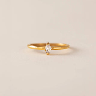 A delicate yellow gold ring featuring a marquise-cut clear gemstone in a prong setting, centered on a warm beige background.