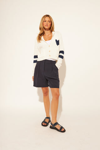 Woman wearing a white cardigan with blue accents, navy shorts, and sandals on a plain background