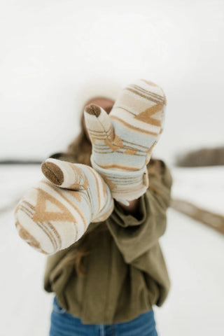 A person in a snowy outdoor setting wearing patterned knit mittens and an olive green jacket, with the mittens held up toward the camera.
