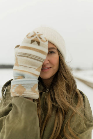 A woman outdoors in a snowy setting wearing a cream knit beanie and a patterned knit mitten over one eye. The mitten has beige, brown, and light blue geometric stripes and motifs.