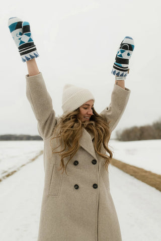 A woman standing outdoors in a snowy field, wearing a beige double-breasted coat, a cream knit beanie, and blue, black, and white patterned knit mittens with her arms raised in the air.