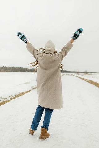 A person standing in a snowy field with arms raised in the air, wearing a long beige coat, blue jeans, tan boots, and blue-and-white striped knit mittens.