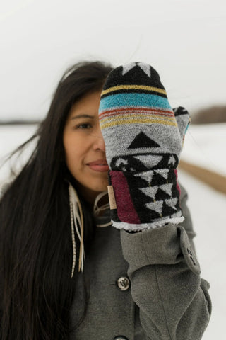 A woman in a gray coat holds up a knit winter mitten with a Southwestern-style geometric pattern in gray, black, turquoise, yellow, and burgundy, set against a snowy outdoor background.