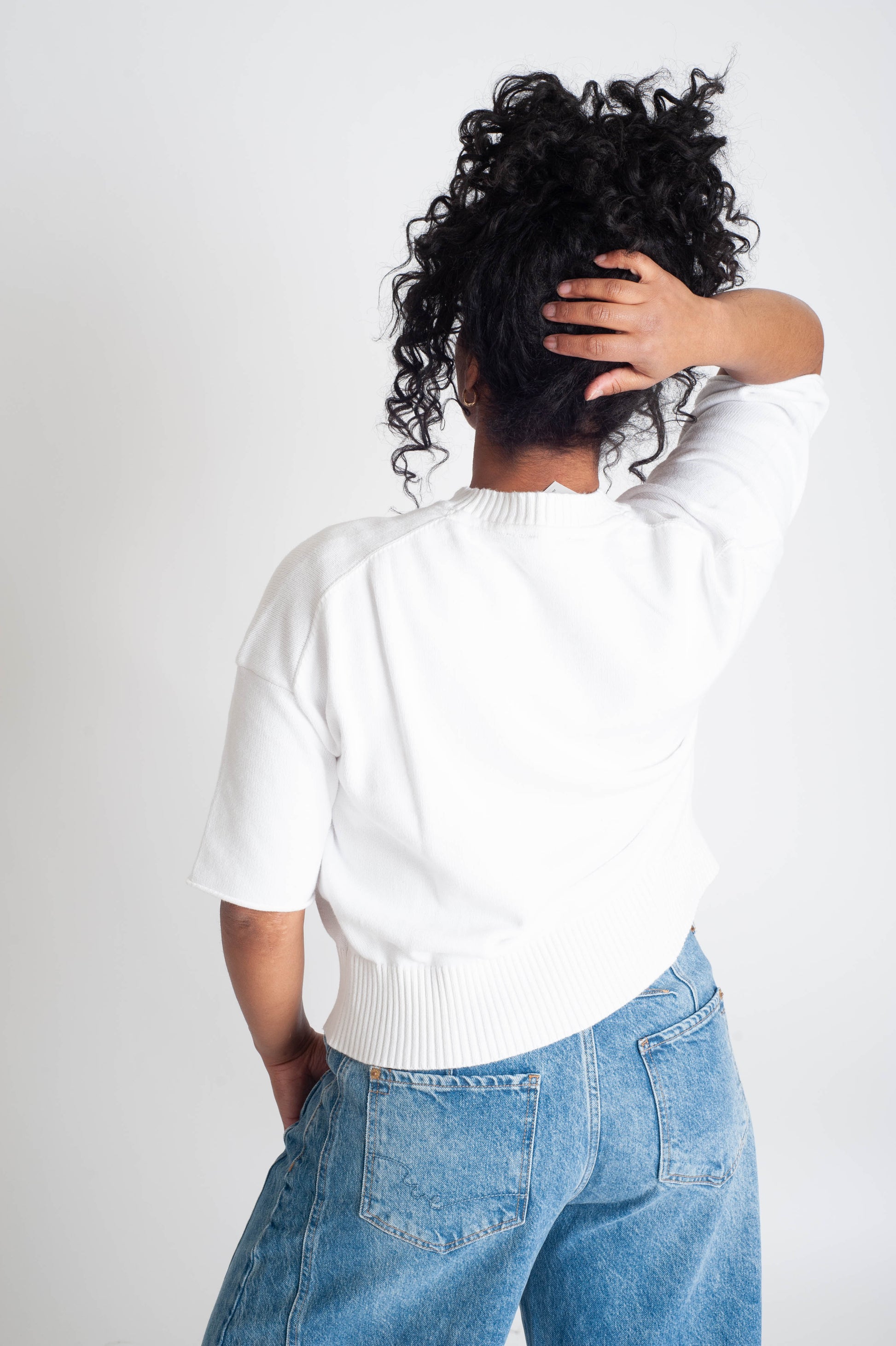 Person wearing a white sweater and blue jeans with hands in hair on a plain background