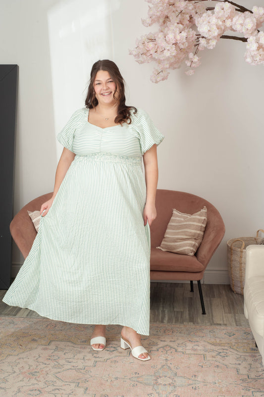 Woman in a light green dress standing in a room with a brown chair and decorative elements.