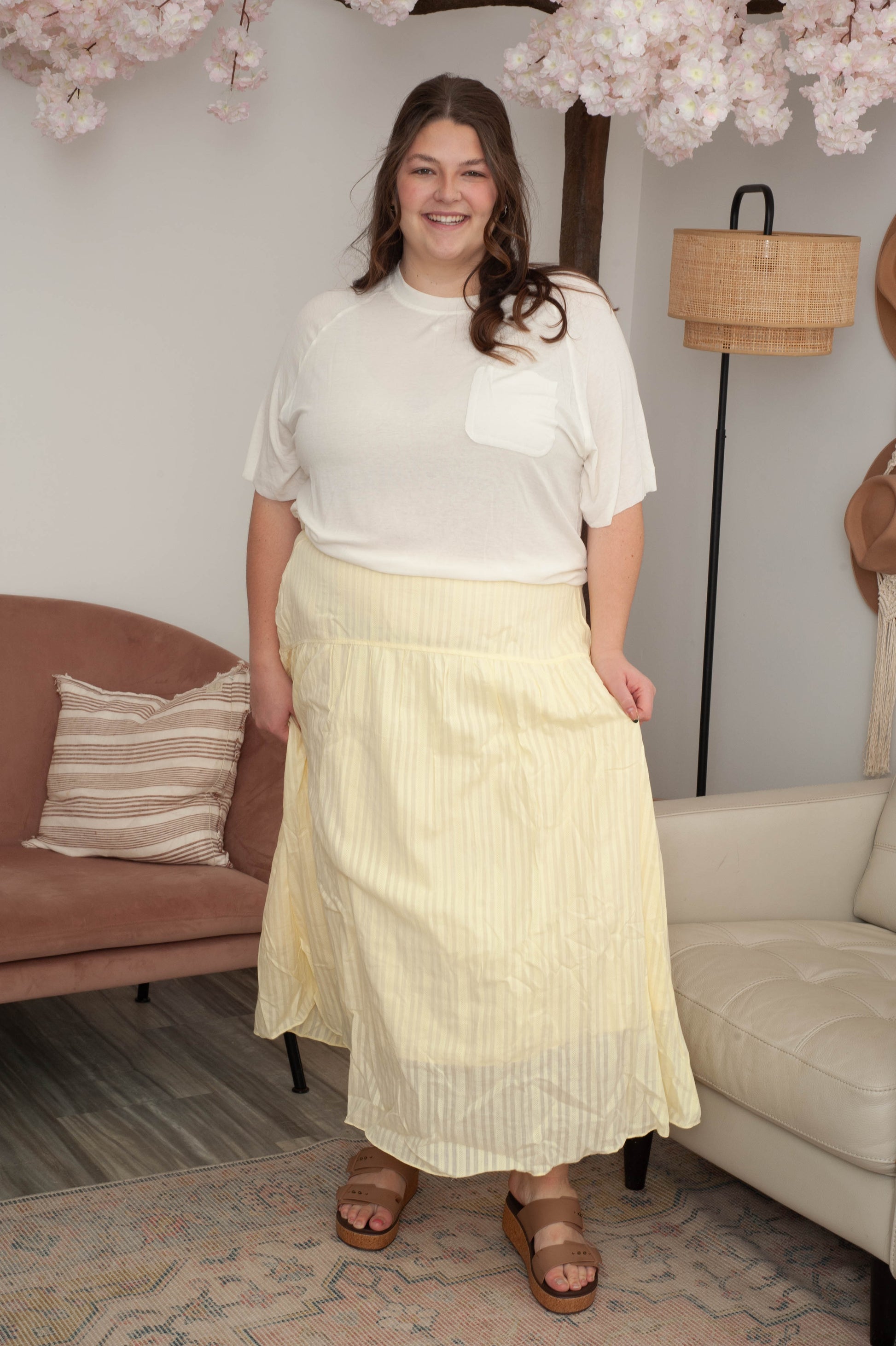 Woman in a white blouse and yellow skirt standing in a living room with a couch and lamp in the background.