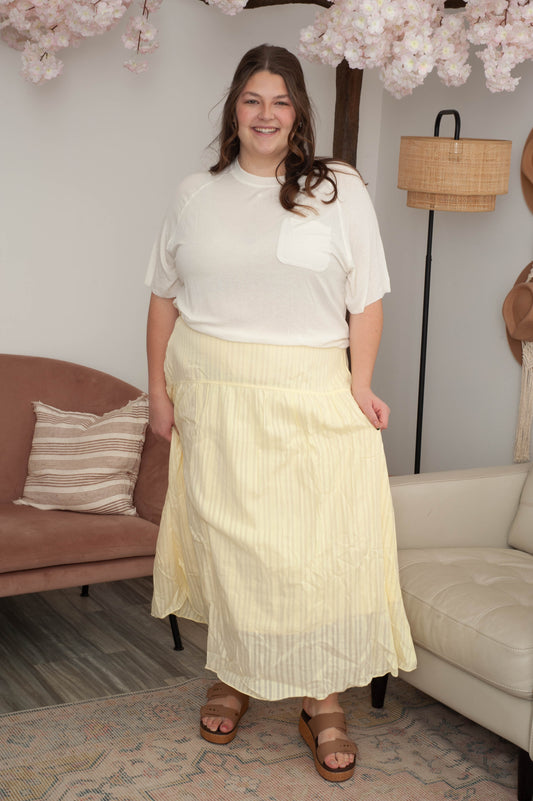 Woman in a white blouse and yellow skirt standing in a living room with a couch and lamp in the background.