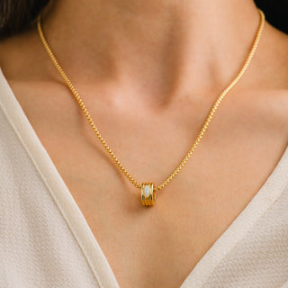 A close-up of a model wearing a delicate gold chain necklace with a small cylindrical pendant featuring a white inlay.