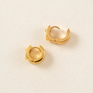 A pair of polished gold crescent-shaped hoop earrings on a light beige textured background.