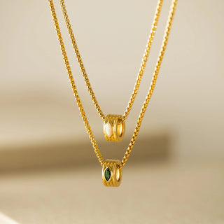Two delicate gold chain necklaces with cylindrical bead pendants, one plain gold and one with a small green marquise-cut stone, displayed against a soft beige background.