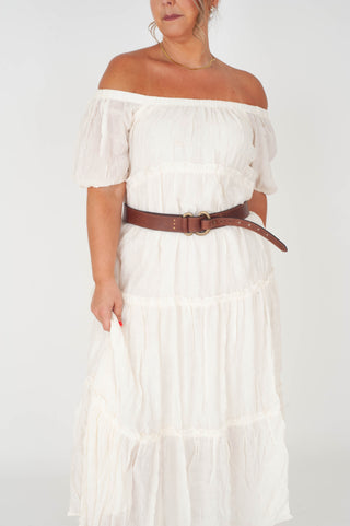 Woman wearing a white off-shoulder dress with a brown belt on a plain background