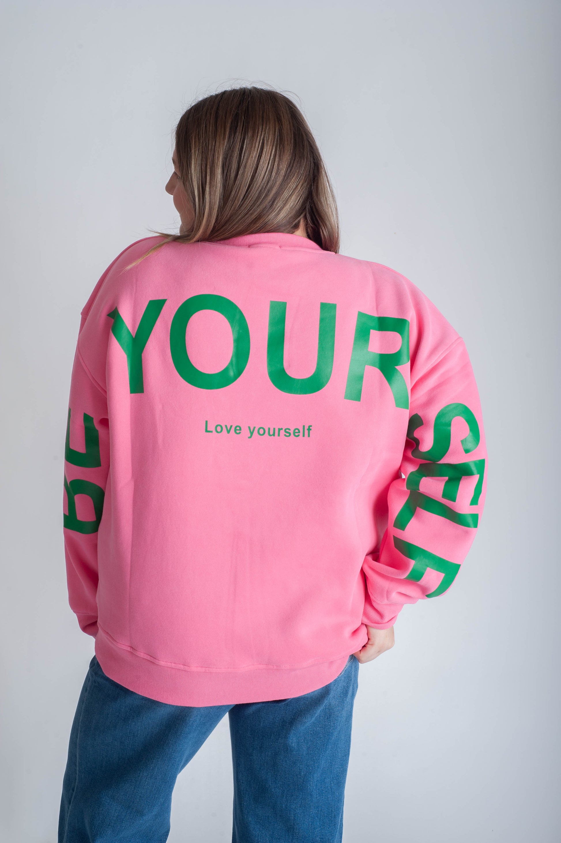 Person wearing a pink sweatshirt with green text on the back, standing against a plain background
