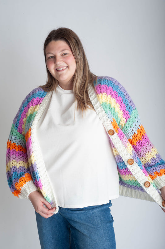 Person wearing a colorful crocheted cardigan with a white shirt and blue jeans on a light gray background
