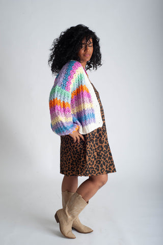 Person wearing a colorful striped sweater, white shirt, leopard print skirt, and beige boots on a plain background
