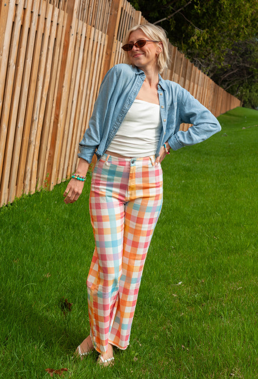 Woman wearing a light blue denim jacket and colorful checkered pants standing on grass with a wooden fence in the background.