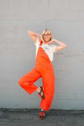 Person wearing bright orange overalls and a white t-shirt against a gray wall.