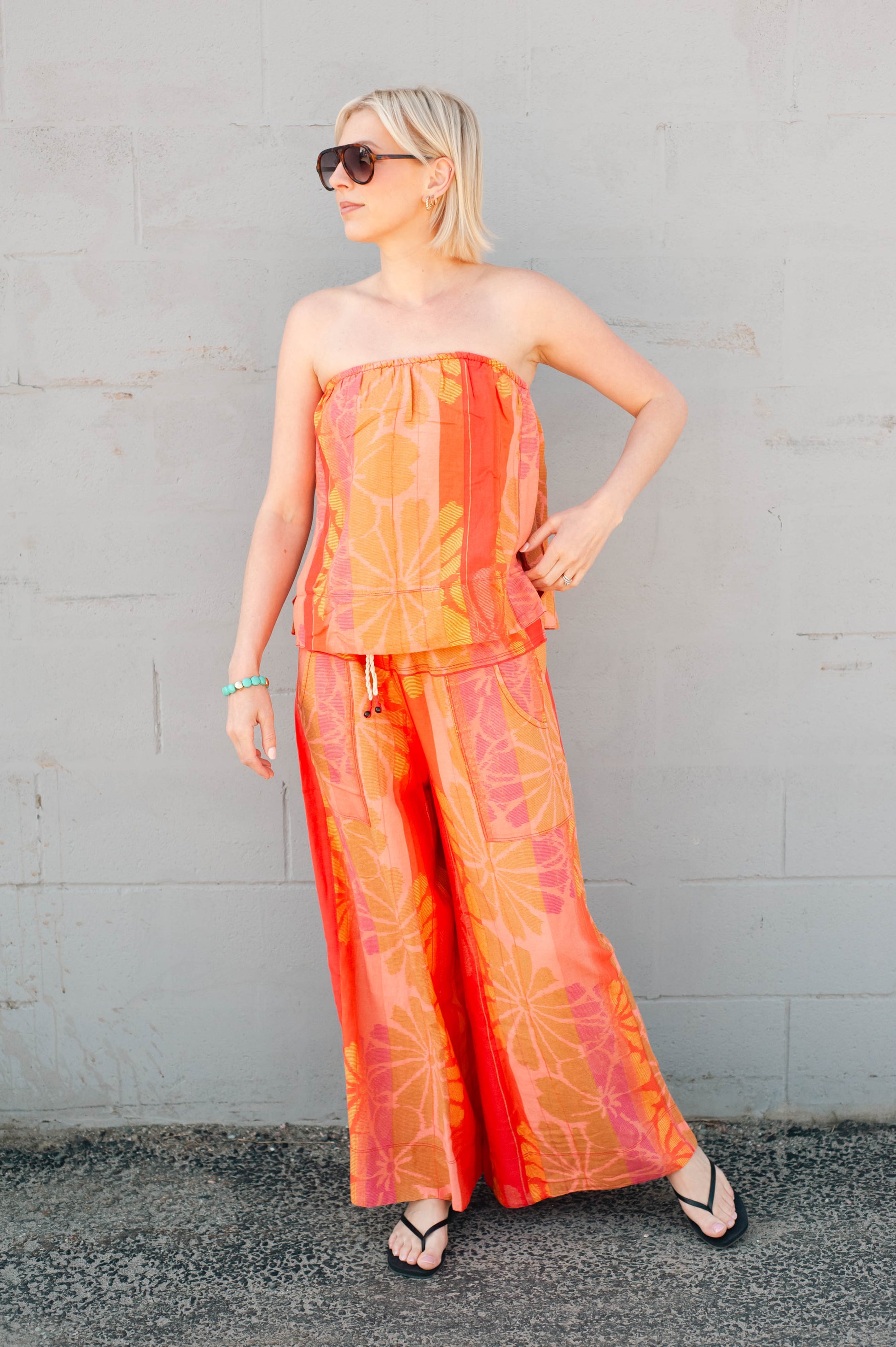 Woman wearing a strapless top and wide-leg pants with a tie-dye pattern against a gray wall.