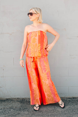 Woman wearing a strapless top and wide-leg pants with a tie-dye pattern against a gray wall.