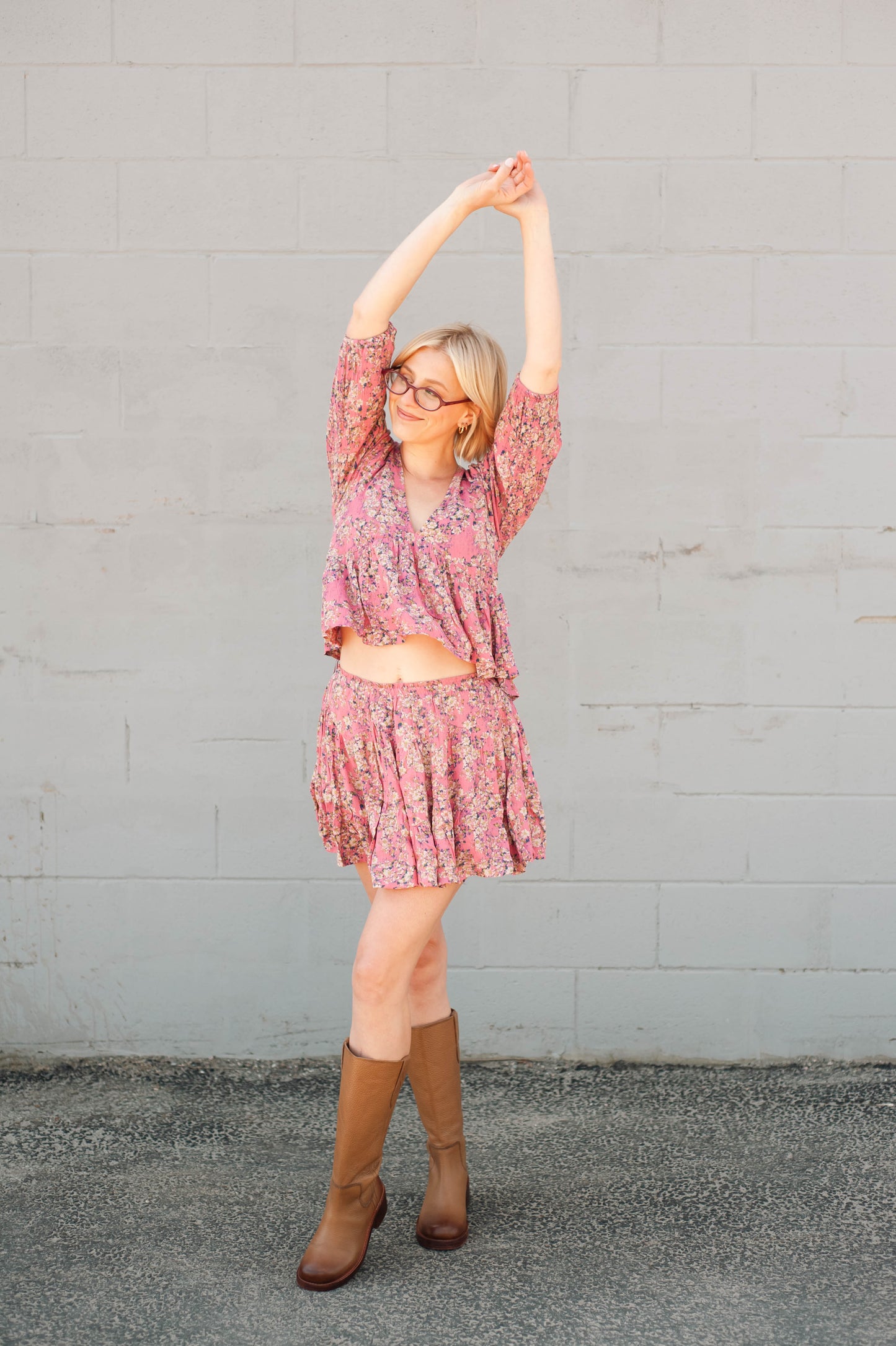 Woman in a pink floral dress and brown boots stretching against a gray wall.