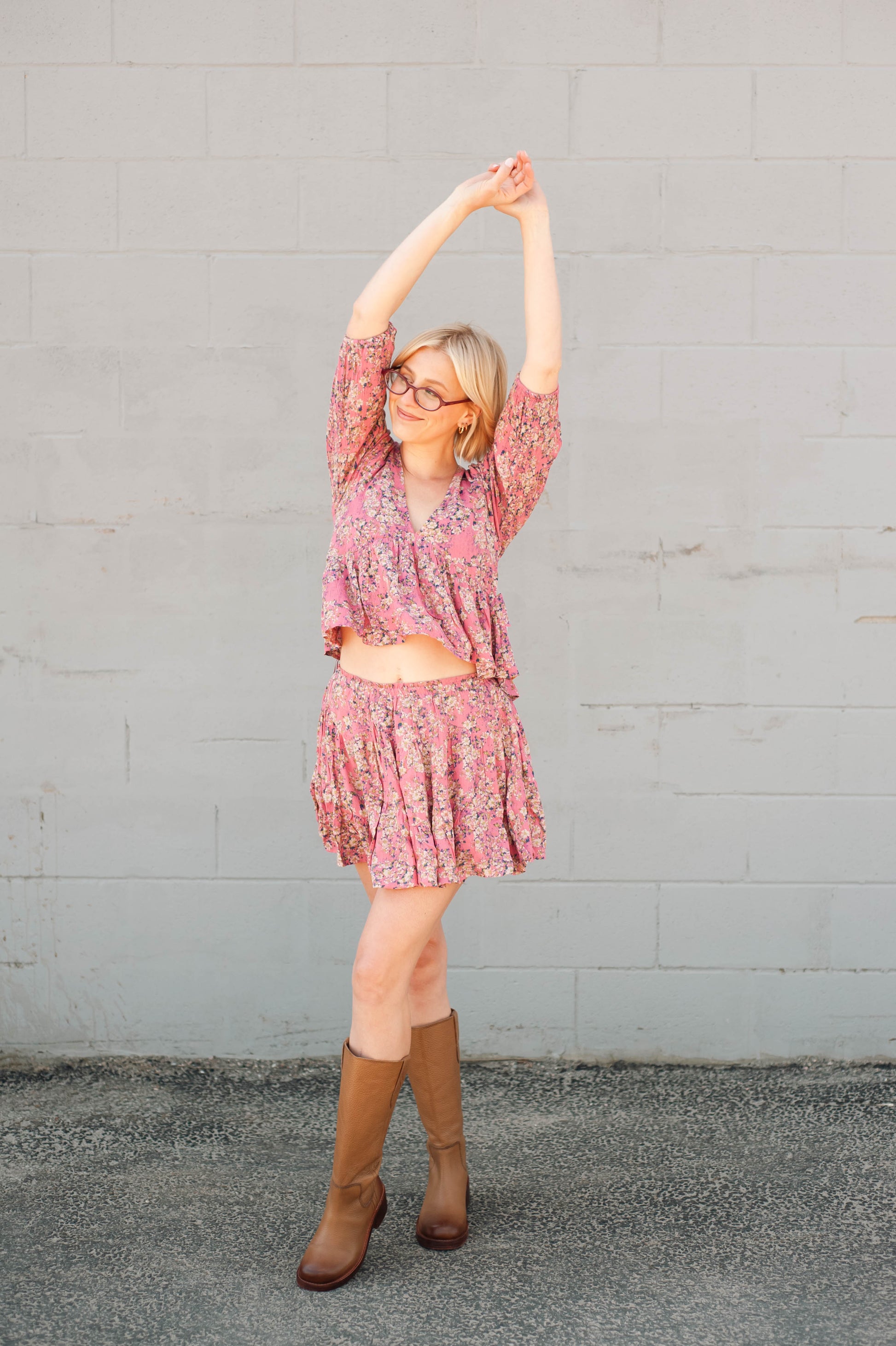Woman in a pink floral dress and brown boots stretching against a gray wall.