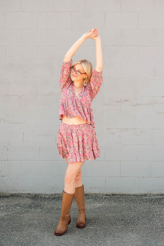 Woman in a pink floral dress and brown boots stretching against a gray wall.
