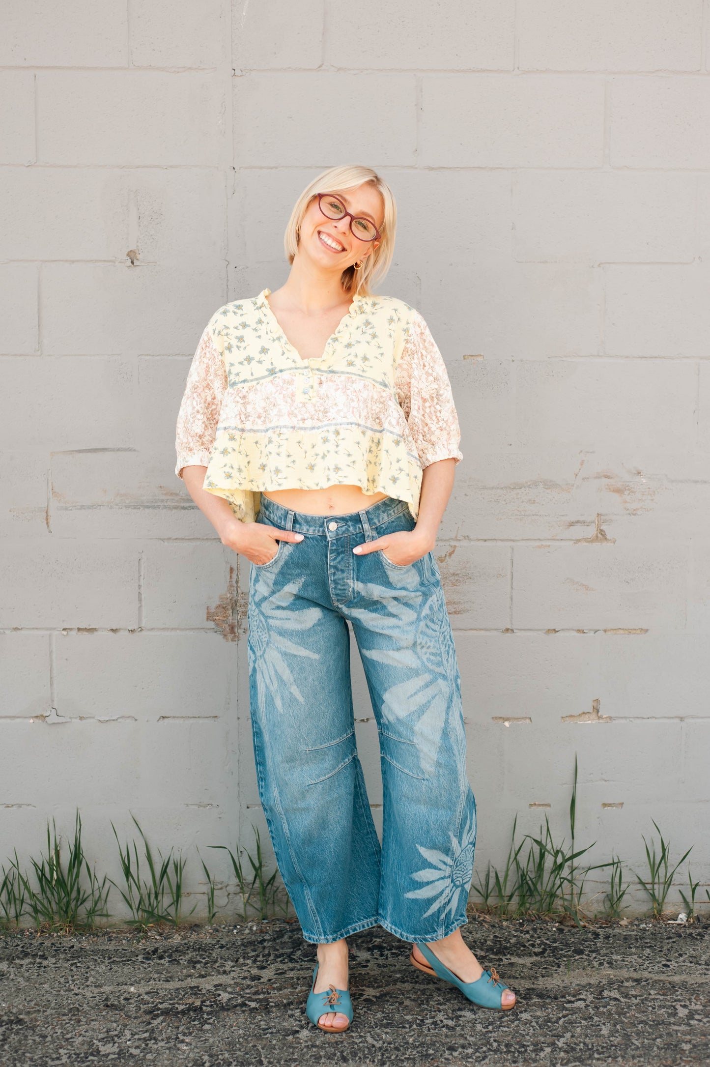 Woman wearing a floral top and blue jeans standing against a gray wall.