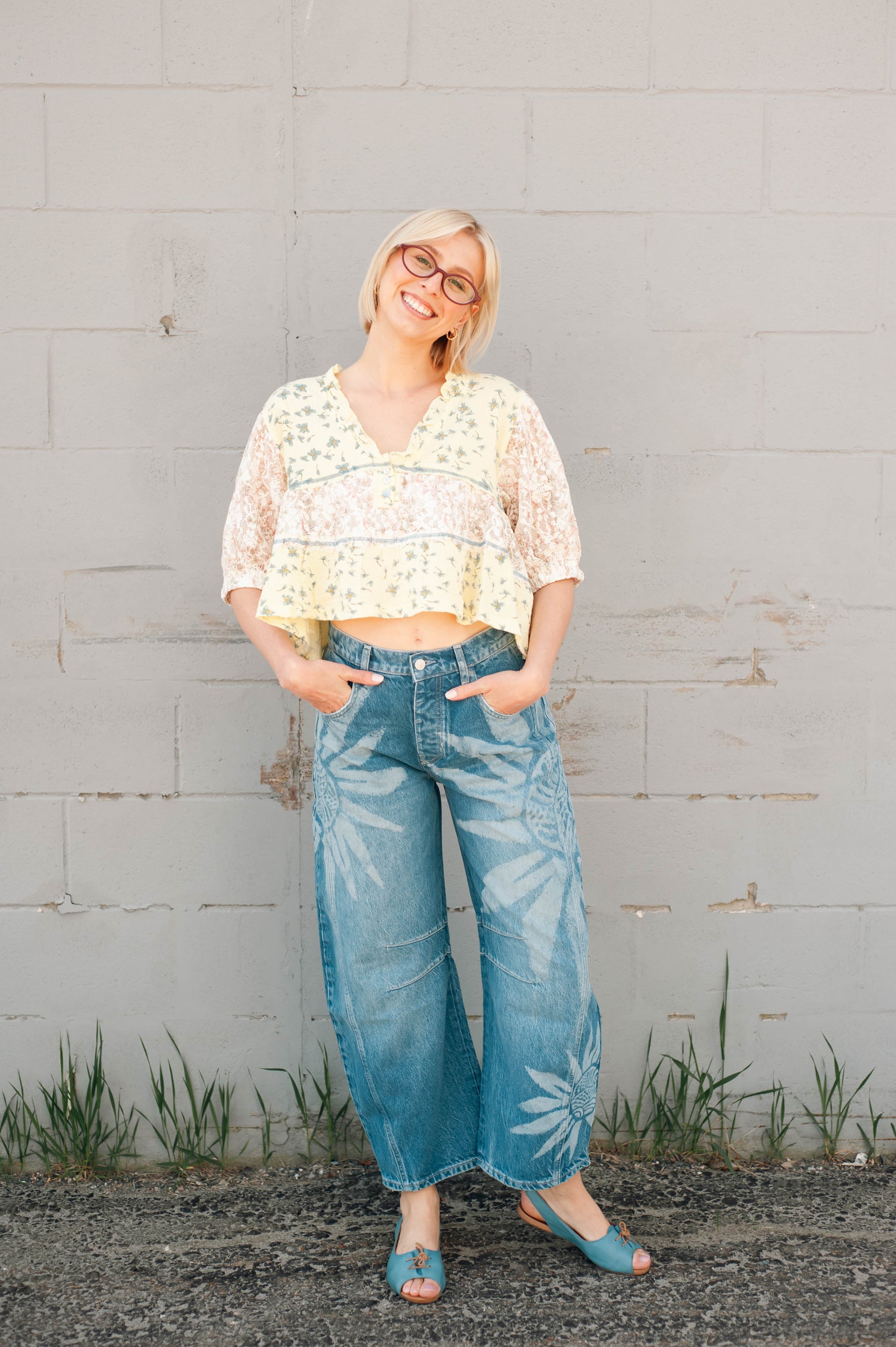 Woman wearing a floral top and blue jeans standing against a gray wall.