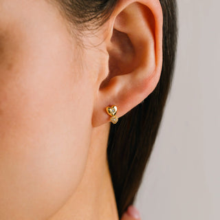 Close-up of an ear wearing a gold heart-shaped earring with a clear gemstone on a neutral background.