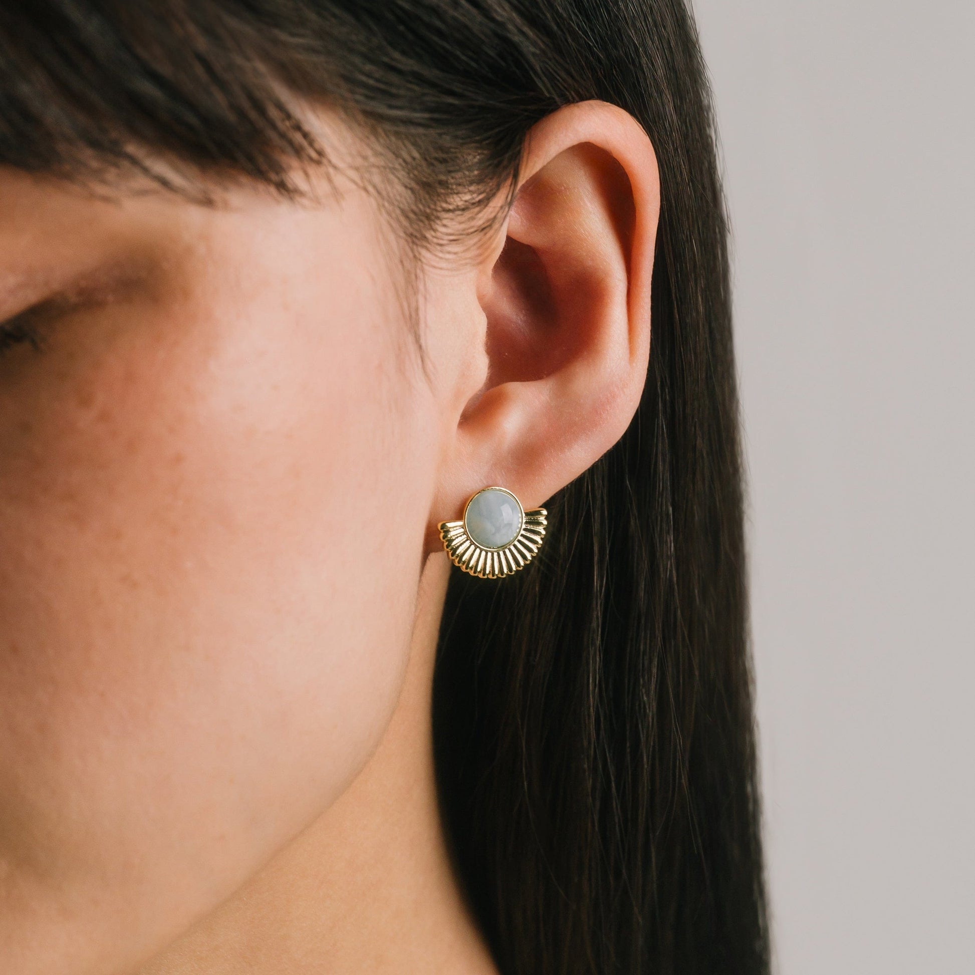 Close-up of an ear wearing a gold earring with a gray gemstone on a neutral background