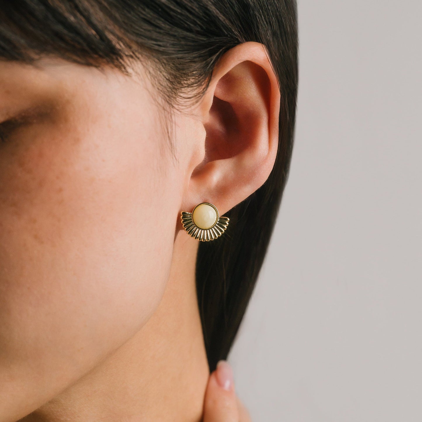 Close-up of an ear wearing a gold earring with a beige center on a neutral background