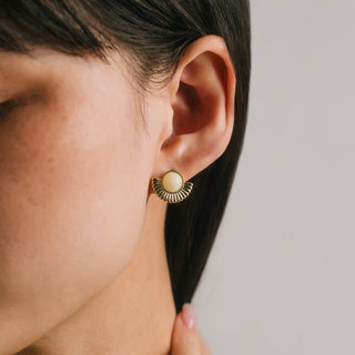 Close-up of an ear wearing a gold earring with a beige center on a neutral background