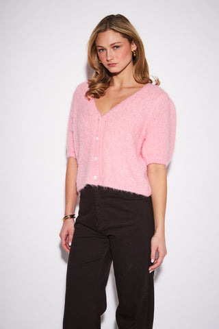 A woman modeling a light pink fuzzy short-sleeve button-up cardigan with black wide-leg pants, standing against a plain white studio background.