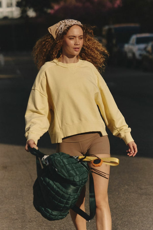Woman in a yellow sweatshirt and brown shorts walking outdoors with a green bag and yellow object.