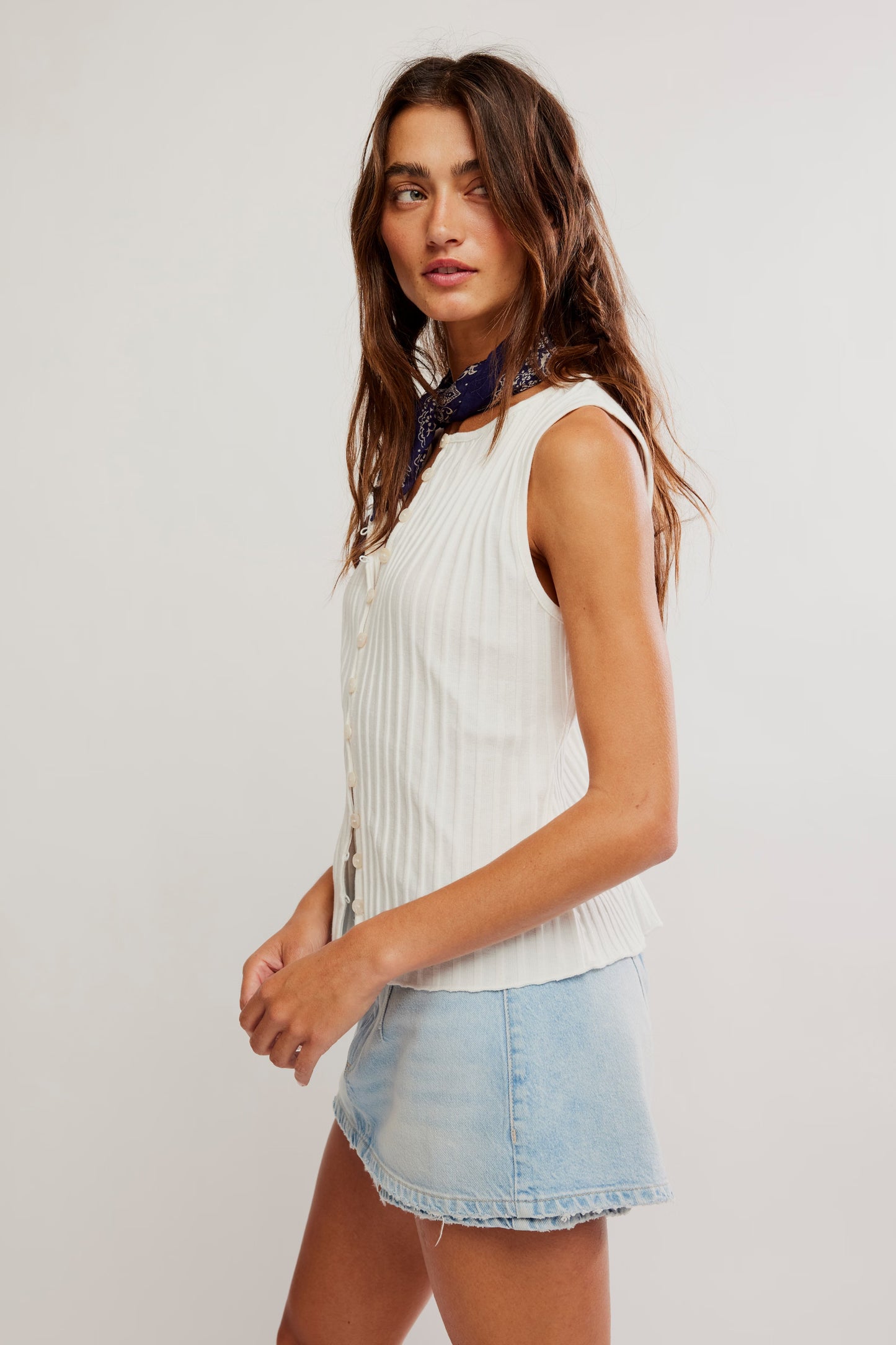 Woman wearing a white sleeveless top and denim shorts with a scarf around her neck on a plain background