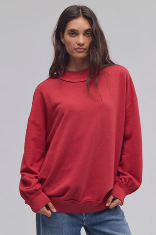 A woman wearing an oversized red crewneck sweatshirt and blue jeans, standing against a solid light gray studio background.