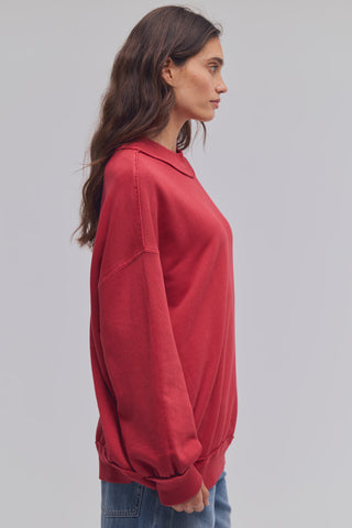 A woman shown in side profile wearing a loose-fitting red long-sleeve sweatshirt and blue jeans against a plain light gray studio background.