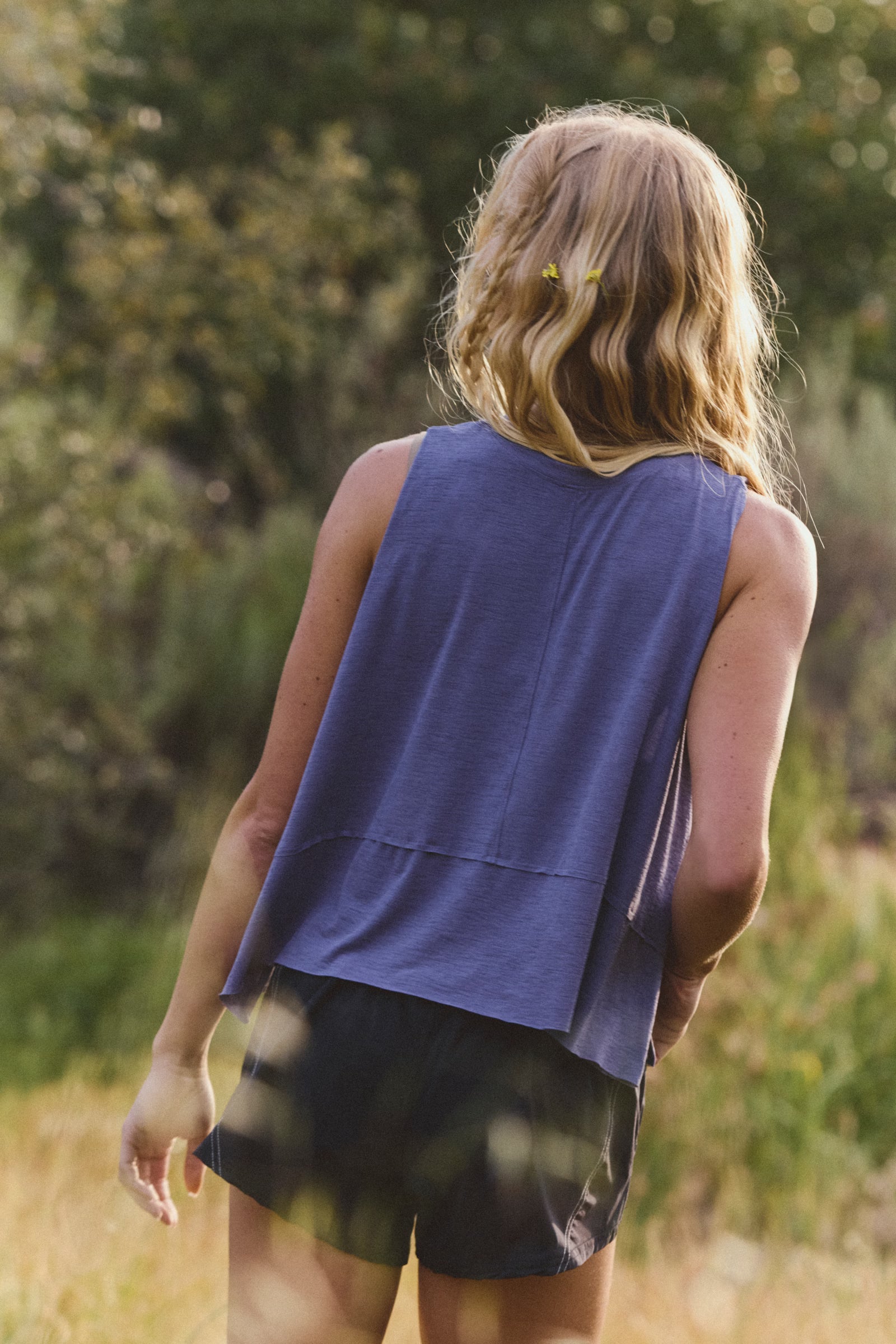 Person wearing a blue sleeveless top and black shorts standing in a natural setting with trees and grass.