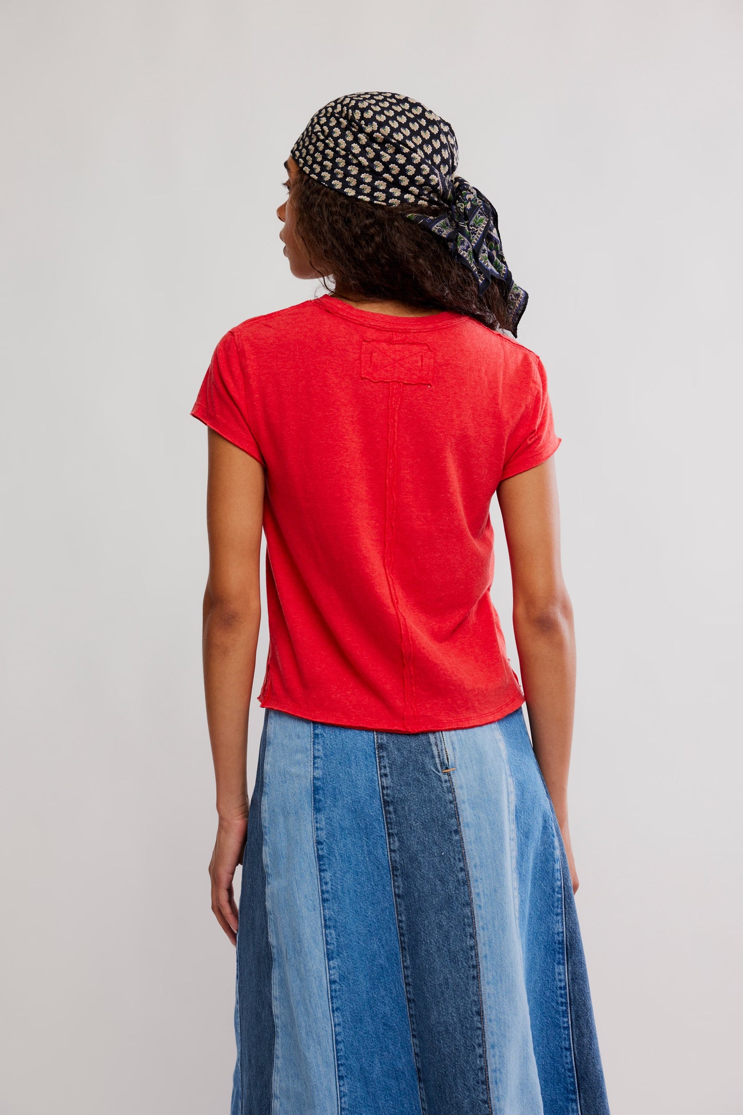 Person wearing a red t-shirt and blue denim skirt with a patterned headscarf on a plain background