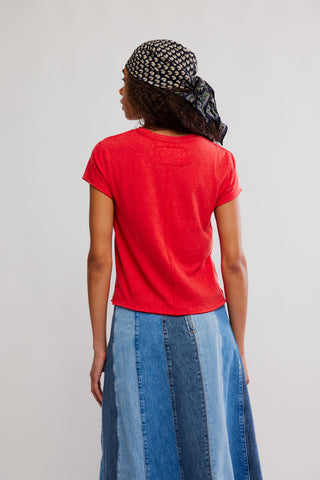 Person wearing a red t-shirt and blue denim skirt with a patterned headscarf on a plain background