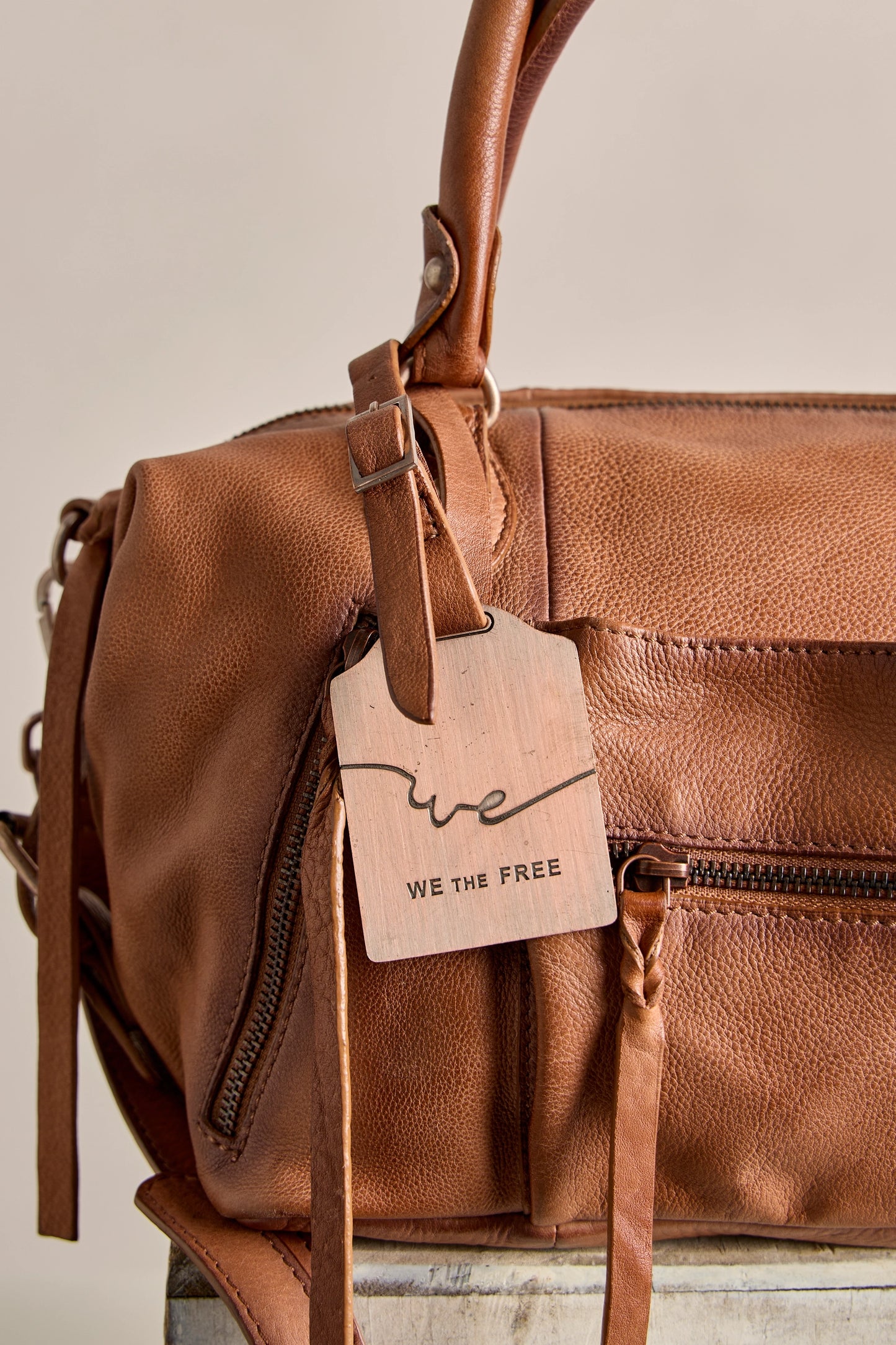 Brown leather bag with a 'WE THE FREE' tag on a neutral background