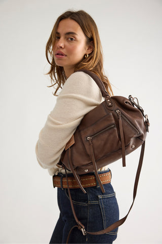 A woman wearing a cream knit sweater and blue jeans carries a brown leather shoulder bag over her shoulder, photographed against a plain light gray studio background.